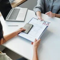 two-people-at-a-desk-exchange-a-clipboard-with-documents-and-a-passport