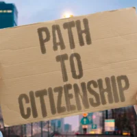 hands-holding-a-cardboard-sign-reading-path-to-citizenship-in-bold-letters-against-a-blurred-urban-backdrop