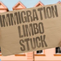 hands-hold-a-cardboard-sign-reading-immigration-limbo-stuck-against-a-backdrop-of-a-building-with-a-redtiled-roof